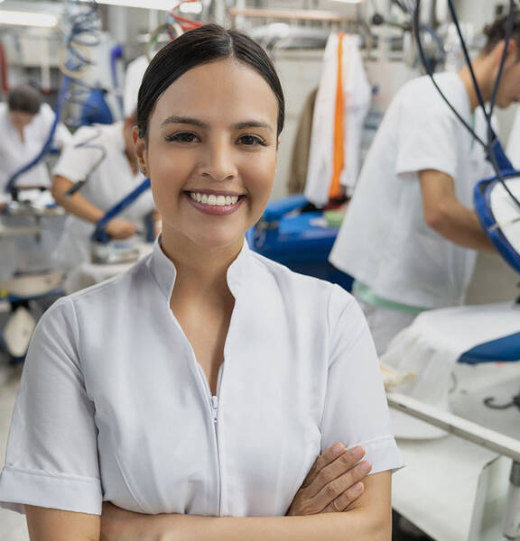 A woman with her arms crossed, smiling as people iron clothes behind her